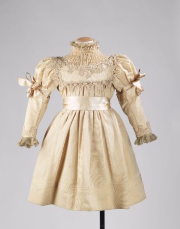 A vintage 1895 girl's party dress with an ivory hue. The dress features intricate lacework around the neckline, sleeves, and bodice, with a smocked high collar. Puff sleeves are adorned with ribbon bows, and the skirt has subtle pleats and embroidery near the hemline.