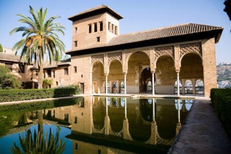 View of a pavilion in the Alhambra Palace, featuring elegant Moorish arches with intricate latticework and a central tower, reflected beautifully in a tranquil rectangular pool. The surrounding greenery, including palm trees and hedges, enhances the serene atmosphere, while the backdrop showcases the hilly landscape of Granada, Spain. The structure highlights the exquisite Islamic architecture of the 14th century.