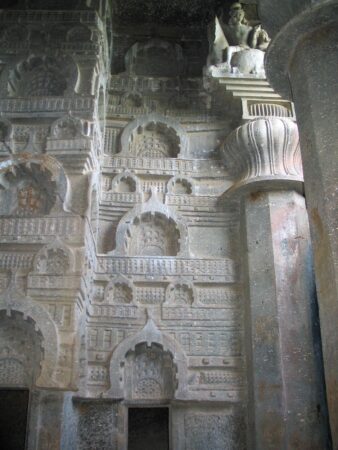 Intricate rock-cut carvings from the Bedsa Caves in Maharashtra, India, dating to the 1st century CE. The wall features a series of ornate chaitya windows, sculpted in repeating patterns, showcasing ancient architectural detailing. A decorative column with a lotus design and a partially visible sculpture of a seated figure are present in the upper corner, highlighting the artistic finesse of the era.