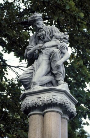 A detailed photograph of a historical statue in Public Gardens near Commonwealth Avenue entrance, Boston, MA, depicting a compassionate figure, likely a healer or doctor, cradling an unconscious or suffering individual. The statue is mounted on a column adorned with carved oak leaves, symbolizing strength and endurance. The figures are sculpted with intricate detail, emphasizing emotion and care. The background features lush green trees, adding a natural setting to this commemorative artwork, likely honoring medical advancements or humanitarian efforts.