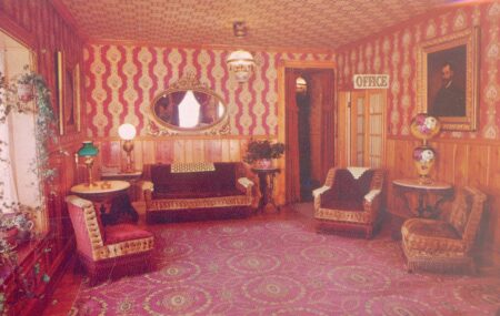 This is a photo of a vintage-style sitting room, featuring rich, ornate decor. The walls are adorned with red and gold patterned wallpaper, and wooden paneling lines the lower half. The room contains upholstered furniture, including chairs and sofas with intricate fringe and fabric details. An oval mirror with a decorative gold frame hangs on the wall, and antique lamps with vibrant colors are placed on side tables. A framed portrait and a sign reading "OFFICE" add to the historical charm of the setting. The rug is bold with circular patterns, complementing the overall Victorian-era aesthetic.