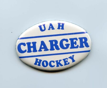 This is a circular button featuring the text "UAH Charger Hockey" in bold blue lettering, with two horizontal blue lines separating the words. The design is set against a white background, likely representing the University of Alabama in Huntsville's hockey team. The button's vintage style suggests it is a memorabilia item, possibly from the 1980s.