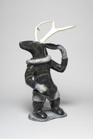 Stone and antler sculpture of a standing humanoid figure with the head of a caribou and full antlers. The figure wears detailed winter clothing, including mitts, boots, and a textured parka trim, and lifts one hand to its hood in a dynamic pose.