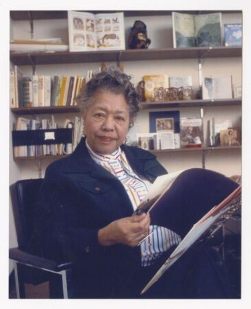 Portrait of Augusta Baker seated in an office chair, holding an open book and looking toward the camera. She is wearing a dark blazer over a striped blouse. Behind her, shelves are lined with children’s books and story props, highlighting her lifelong work in children’s literature and storytelling.
