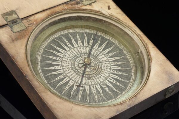 A 17th-century magnetic compass housed in a wooden case with brass hinges. The compass features a detailed wind rose with cardinal directions and degree markings engraved on a circular plate. The needle points north, and the ornate design includes concentric rings of numbers and letters used for navigation.